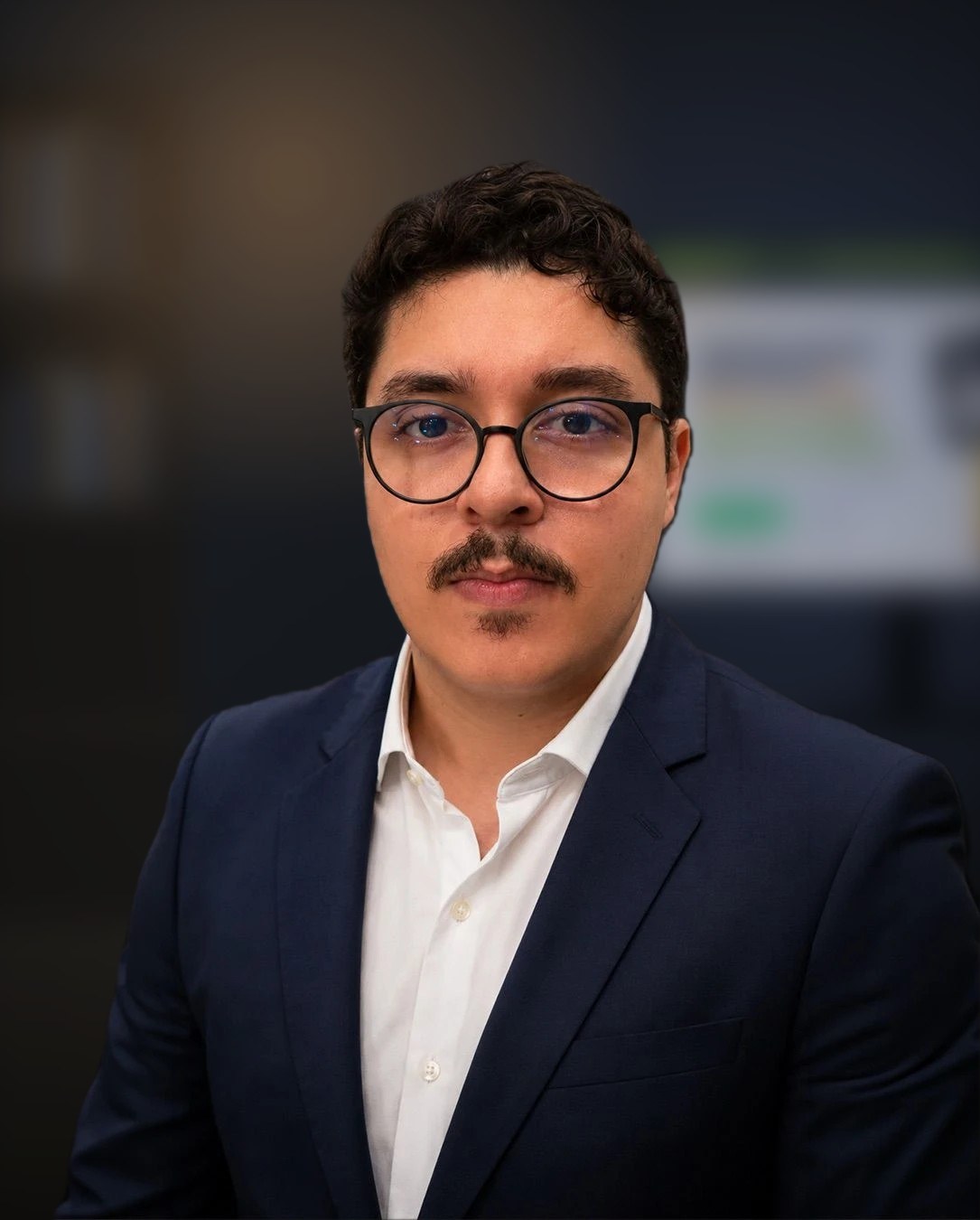 Pedro Cavalcant de óculos, camisa branca e paletó azul-marinho, em um escritório com um monitor ao fundo